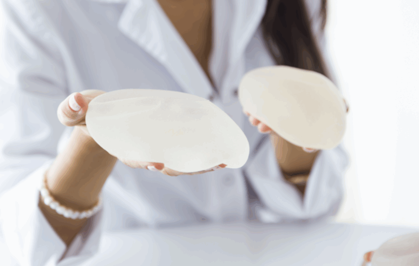 一位穿著白袍和珍珠手鍊的人，雙手各展示一個光滑的乳房植入物