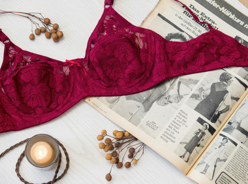 一件深紅色蕾絲胸罩平鋪雜誌上，旁邊裝飾有燃燒的蠟燭和乾枯的漿果枝，呈現女性內衣、時尚或復古風格的靜物攝影