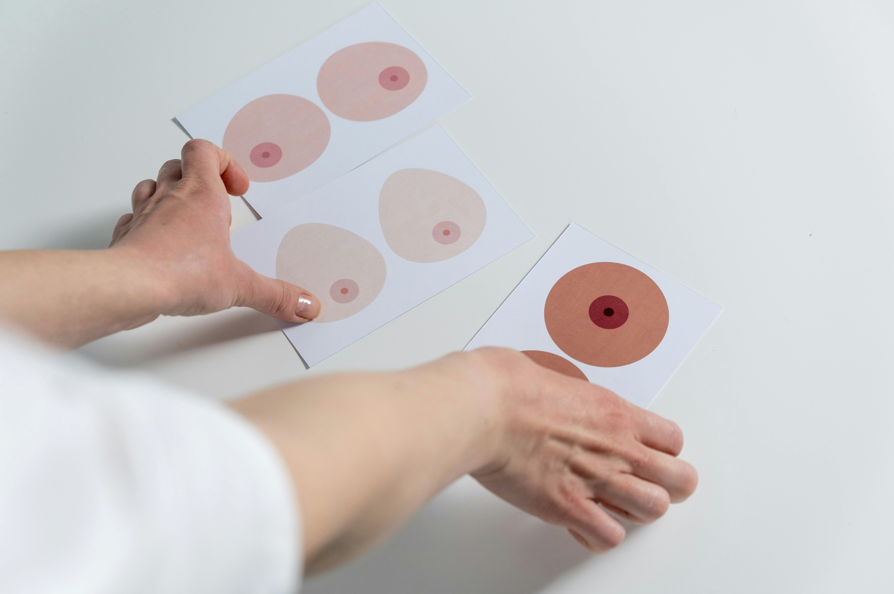 一雙手正在擺放三張卡片或紙張，上面印有不同膚色和形狀的乳房與乳頭的簡化圖案，用於乳房健康教育意識宣傳
