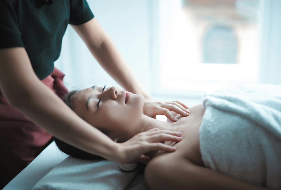一名女性顧客臉上帶著放鬆的微笑躺在按摩床上，身上覆蓋著毛巾，按摩師正在輕柔地按摩她的鎖骨和胸部上方區域，突顯放鬆按摩服務。