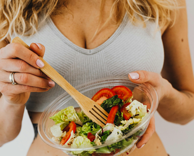 一位穿著運動內衣和運動長褲、身材健美的女性，手拿木製叉子，捧著一碗新鮮的蔬菜沙拉，展現健康飲食生活方式