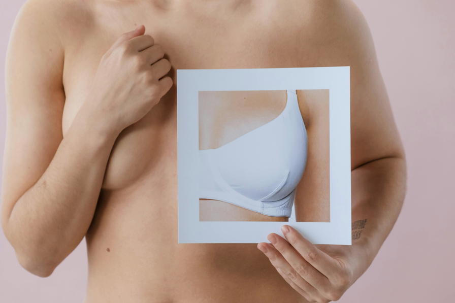 一位上半身裸露的女性，將一張印有白色胸罩或運動內衣照片的紙張拿在胸前。這可能象徵著穿著、乳房健康或乳房美學的主題