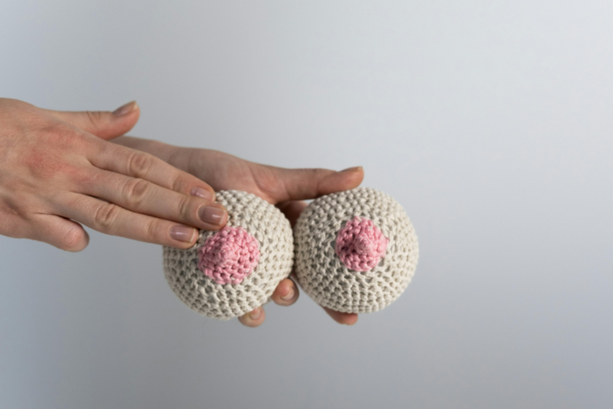 一雙手拿著兩個手工編織的毛線乳房模型，米白色乳房與粉紅色乳頭，常用於乳房檢查衛教