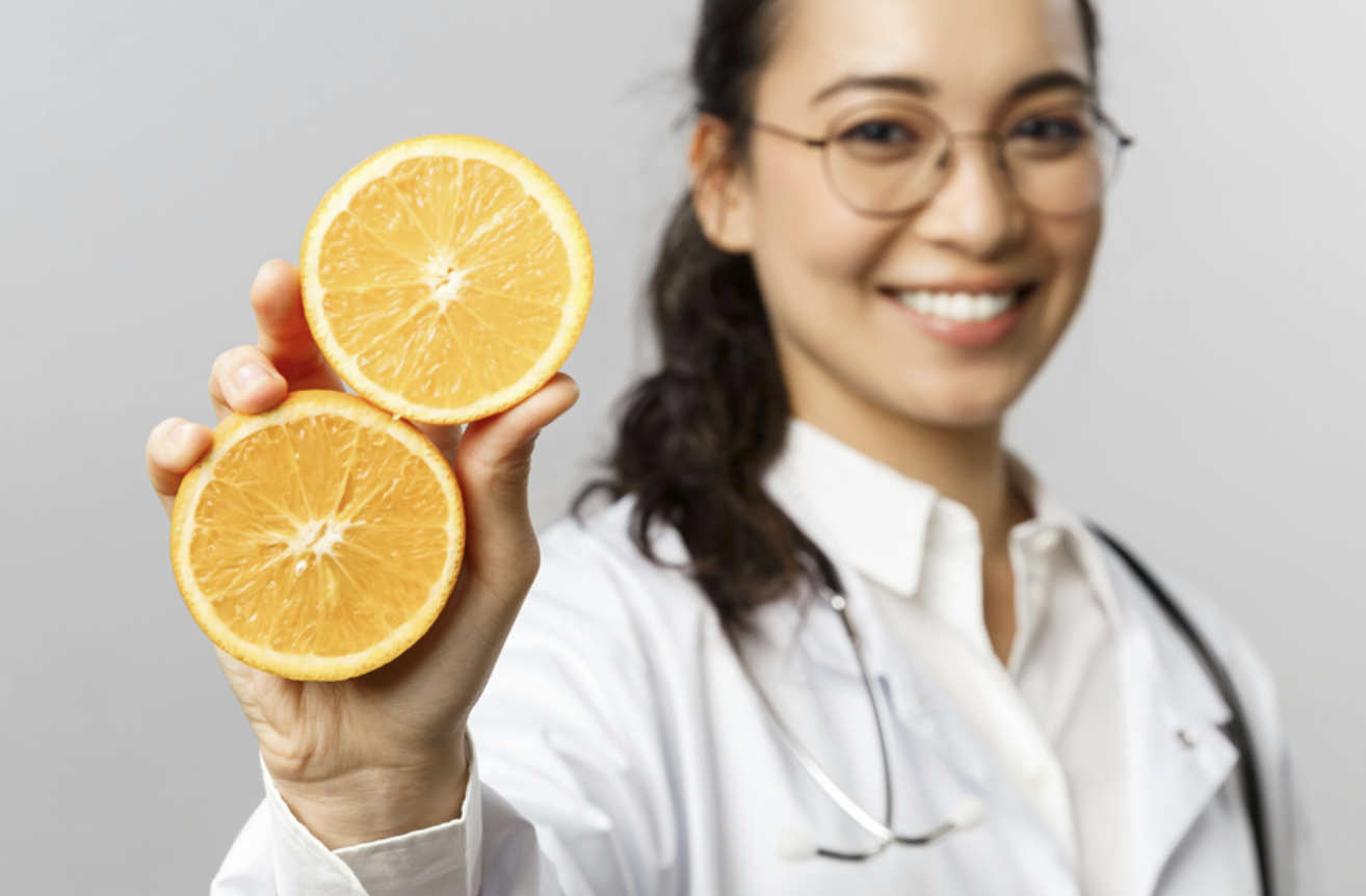 一位戴眼鏡、穿著醫師袍和聽診器的營養師，面帶微笑地伸出手，展示兩片對半切開的橙色柳丁或柑橘類水果，象徵健康飲食