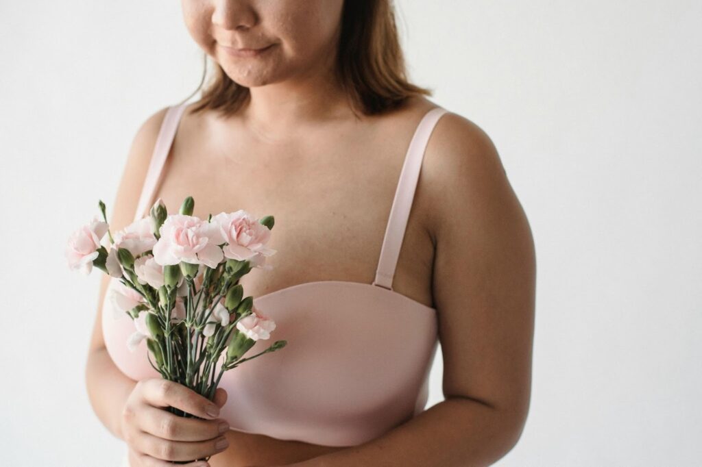 一位女性穿著粉紅色無鋼圈或舒適型胸罩，手中捧著一束粉紅色康乃馨或小花，面帶微笑，象徵乳房健康意識、女性關懷與自信回復。