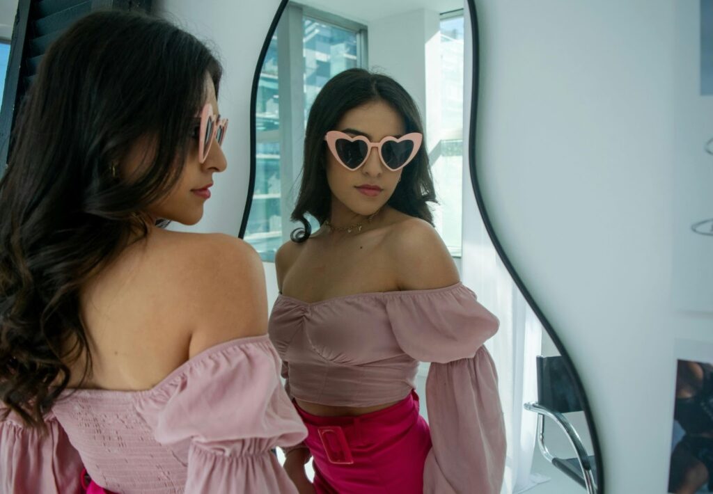 一位穿著粉色露肩上衣和桃紅色褲子的女性，戴著粉色愛心形狀的太陽眼鏡，在鏡子前自拍或審視自己，展現時尚、自信與產後恢復的自然美。