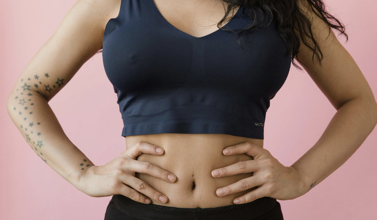 一名手臂上有星星紋身的女性，穿著深藍色運動內衣與黑色長褲，雙手叉腰站在粉紅色背景前