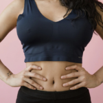 一名手臂上有星星紋身的女性，穿著深藍色運動內衣與黑色長褲，雙手叉腰站在粉紅色背景前