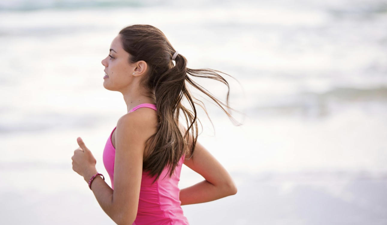 一名綁著馬尾的女性穿著桃紅色運動背心，在海灘上慢跑的側面影像