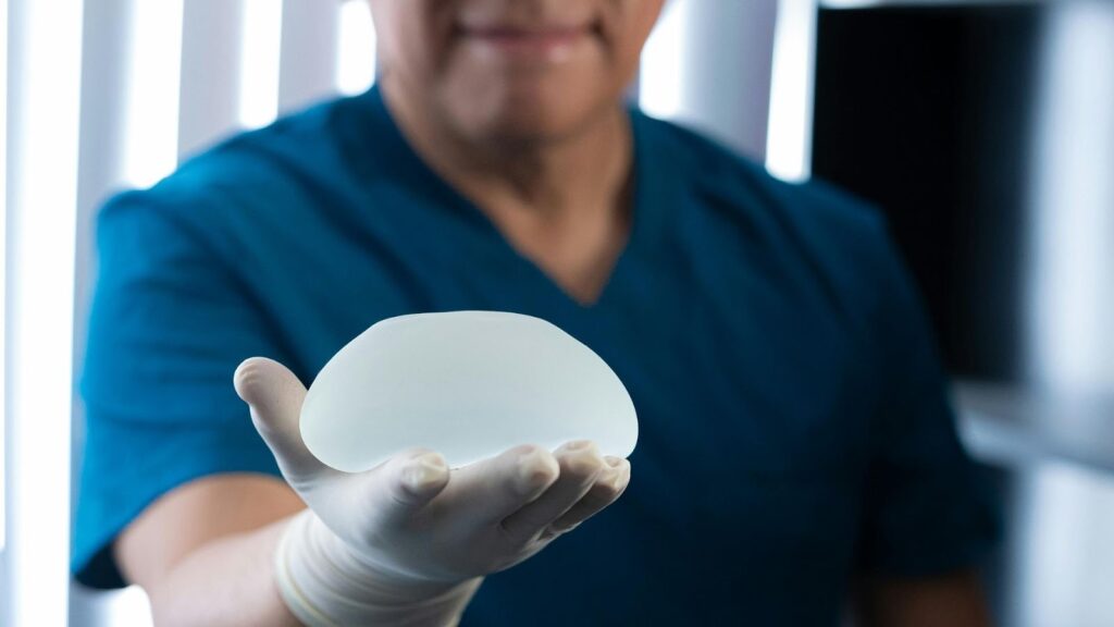一名穿著藍色刷手服的醫療人員或醫師，戴著白色實驗手套，掌心托著一個水滴形的半透明矽膠乳房植體。這張照片以專業的視角呈現了隆乳手術中使用的義乳材質與形狀，適合用於整形外科手術說明或衛教內容。