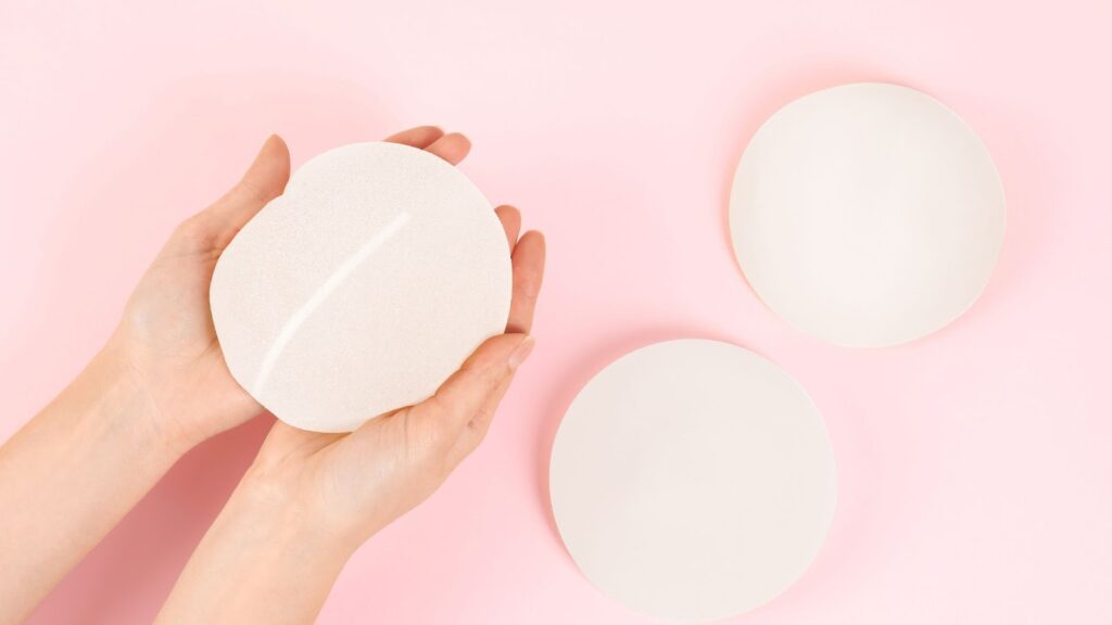 在淡粉色背景上，雙手托著一個圓形矽膠隆乳植入物，旁邊另外擺放著兩個不同規格的植入物，呈現醫學美容中義體選擇與質地展示的場景。