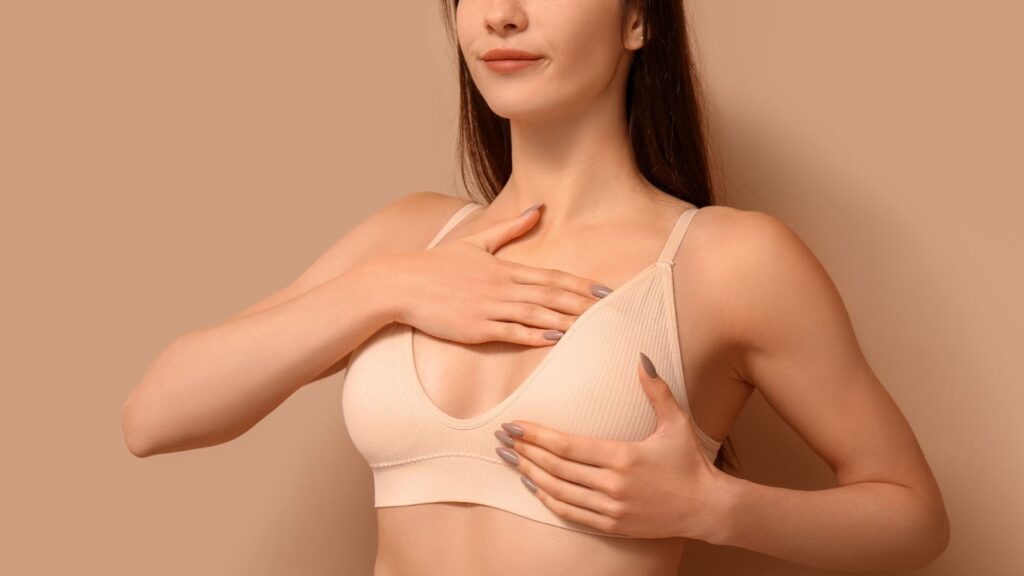一名身穿米色運動內衣或術後壓力衣的女性，雙手輕柔地放置於胸部位置，呈現自我檢查或術後照護的示意畫面。背景色調溫潤，旨在傳達女性健康、隆乳術後保養或乳房健康篩檢的衛教主題。