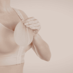 一名穿著膚色運動內衣或術後壓力衣的女性，手中拿著一個半透明的矽膠乳房植體。畫面呈現乾淨且專業的色調，常用於說明隆乳手術、乳房重建或義乳選擇等醫學美容與健康護理情境。