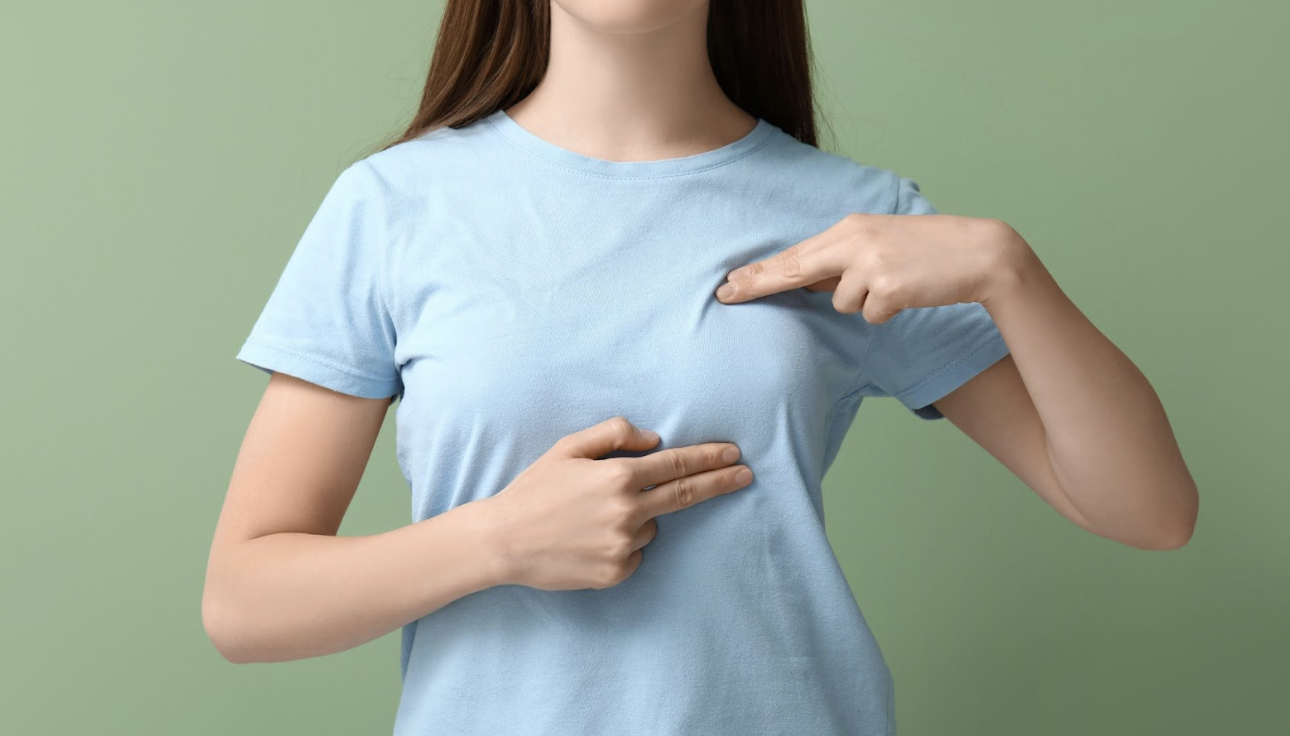 一名穿著淺藍色 T 恤的女性，使用雙手手指指向胸部特定位置，示意自我檢查或諮詢手術改善部位的情境